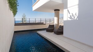 Un espace piscine moderne offrant une vue dégagée sur le paysage urbain, un mur blanc orné d’une plante suspendue et deux chaises longues.