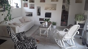 Un salon comprenant un fauteuil à motifs zébrés, un fauteuil à bascule blanc, un canapé blanc et une table basse. Des étagères ornées d’objets décoratifs et de plantes sont fixées aux murs.