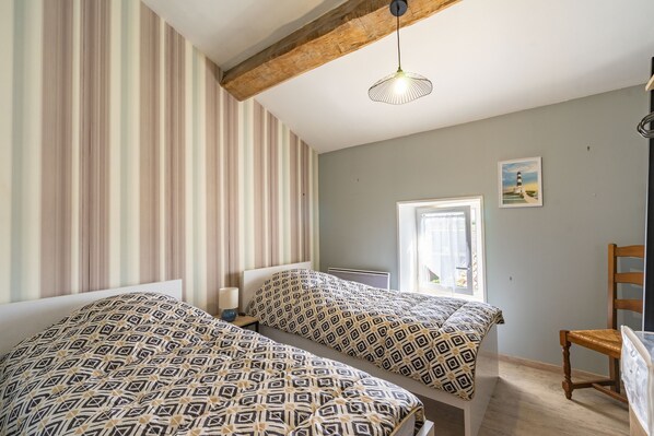 A room with two beds, a wooden ceiling beam, a hanging light fixture, a window with curtains, and a framed picture on the wall.