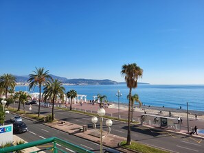 A coastal cityscape with a clear view of the sea, palm trees, and a pedestrian promenade.