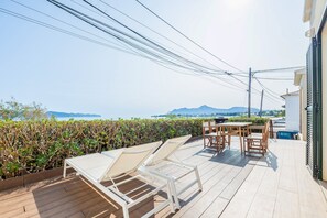Una terrazza in legno con poltrone relax e un tavolo, con vista su un corpo d'acqua e montagne.