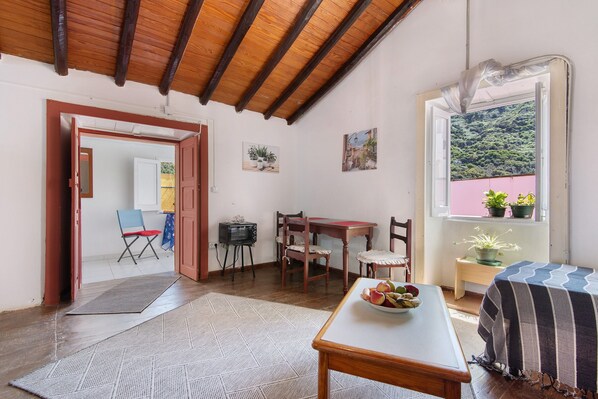 A cozy room with a wooden ceiling, a dining table with chairs, a small TV, and a window with a view of greenery.