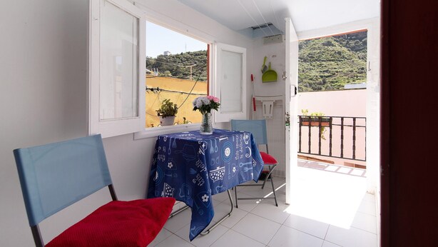 A balcony with a table and chairs, a window with a view of a hill, and a door. - Vacation Home 'Mi Idilio' with Mountain View and Wi-Fi (Teror)