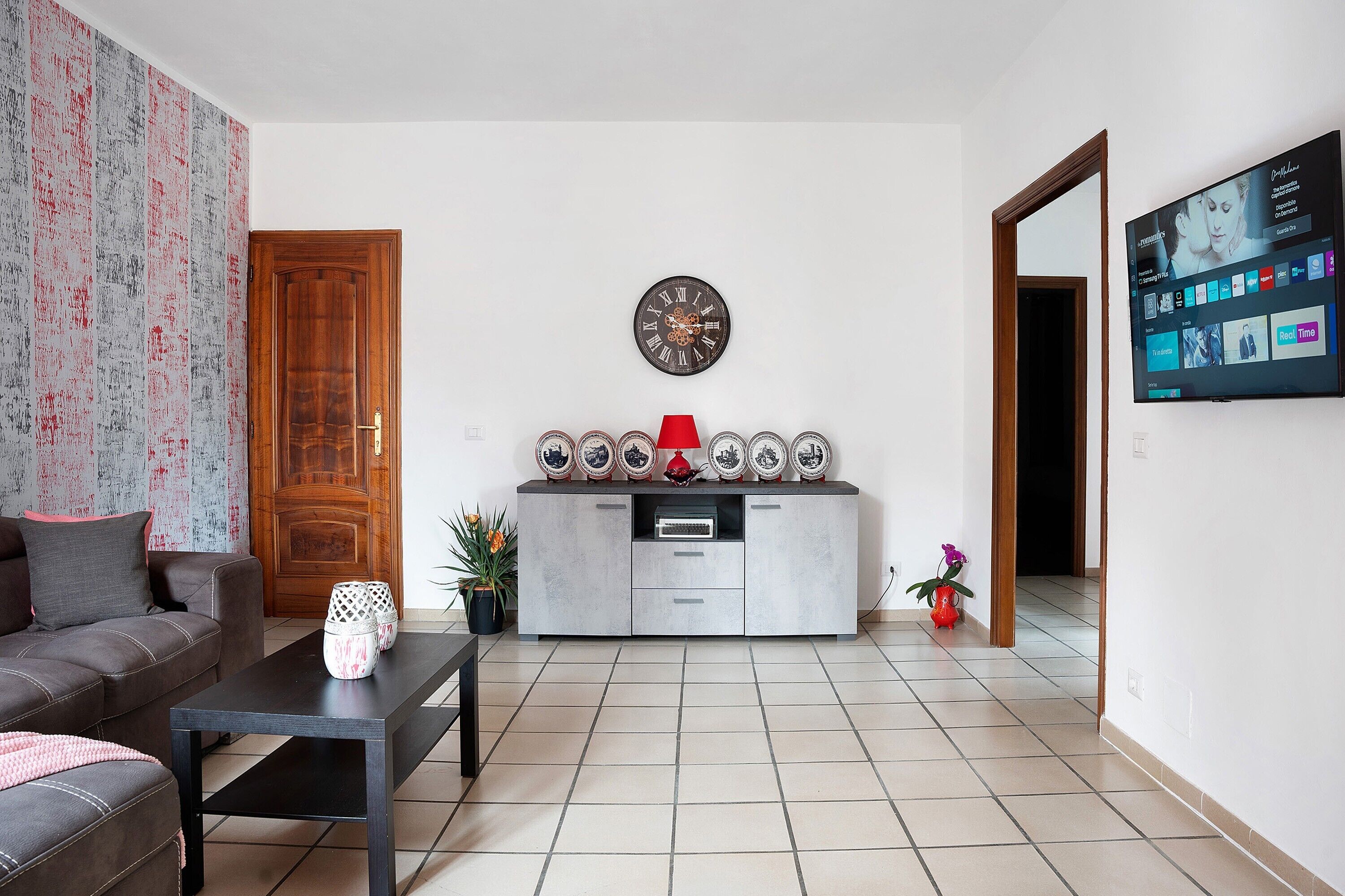 Un salon comprenant un canapé, une table basse, un meuble avec des assiettes décoratives, une horloge et un téléviseur fixé au mur.