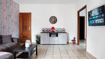 Un salon comprenant un canapé, une table basse, un meuble avec des assiettes décoratives, une horloge et un téléviseur fixé au mur.