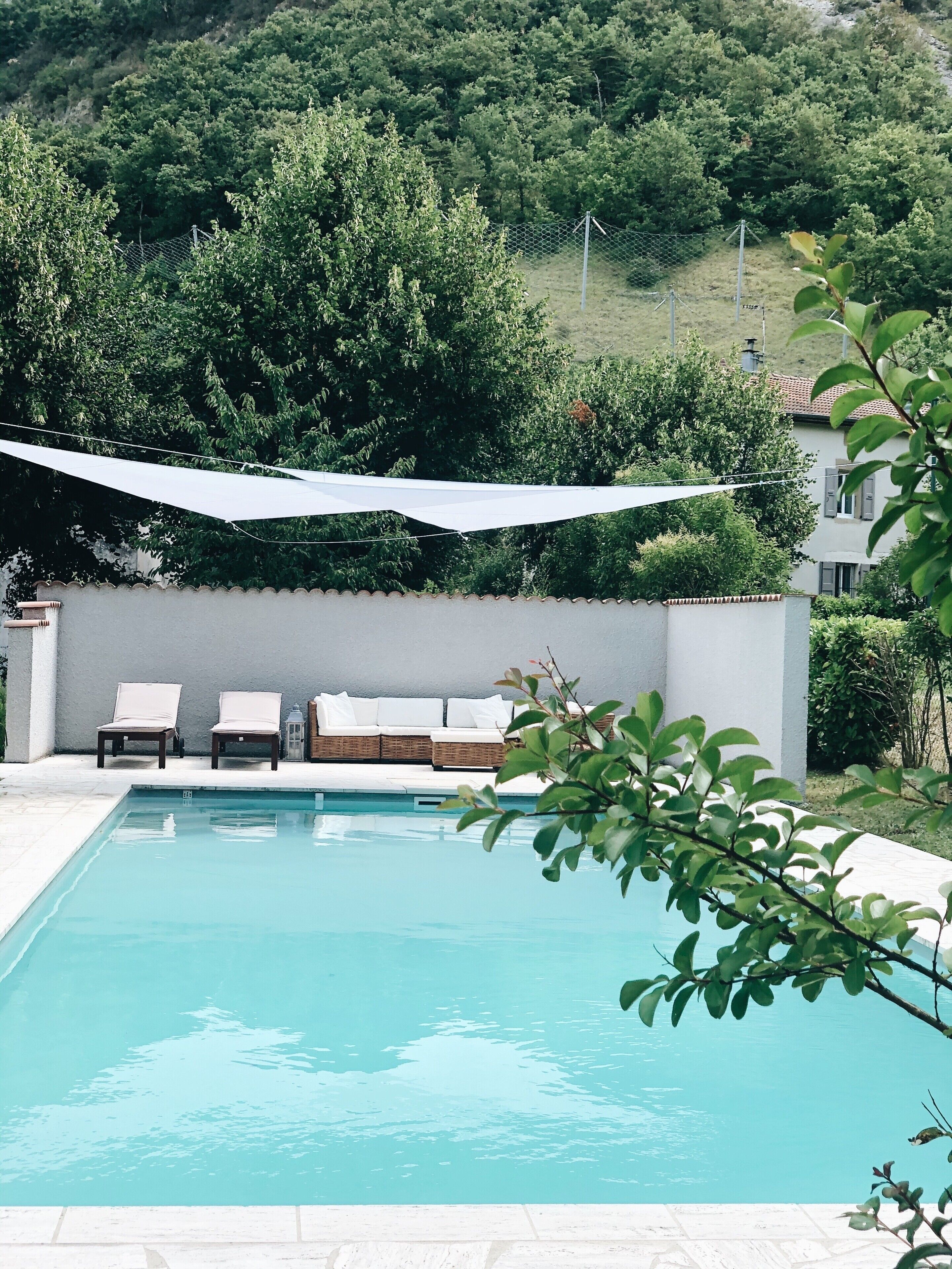 Une piscine rectangulaire dotée d’une terrasse blanche, entourée d’un mur blanc et d’une clôture en treillis métallique. On y trouve des chaises longues et un canapé en osier sous un espace ombragé.