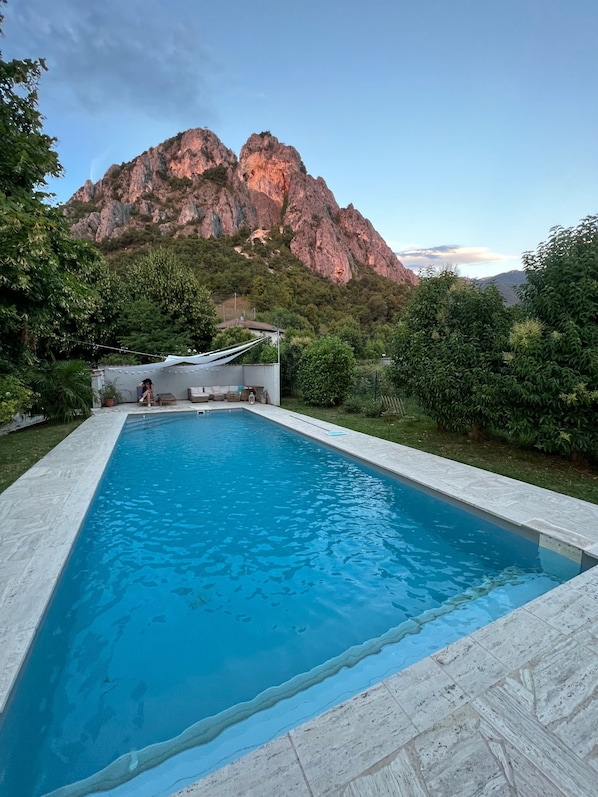 A rectangular swimming pool with clear blue water, surrounded by a white tiled deck. In the background, there is a mountainous landscape with rocky outcrops and greenery.
