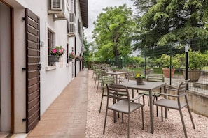 An outdoor patio with tables and chairs, a white building with shutters, and a tree-lined background.