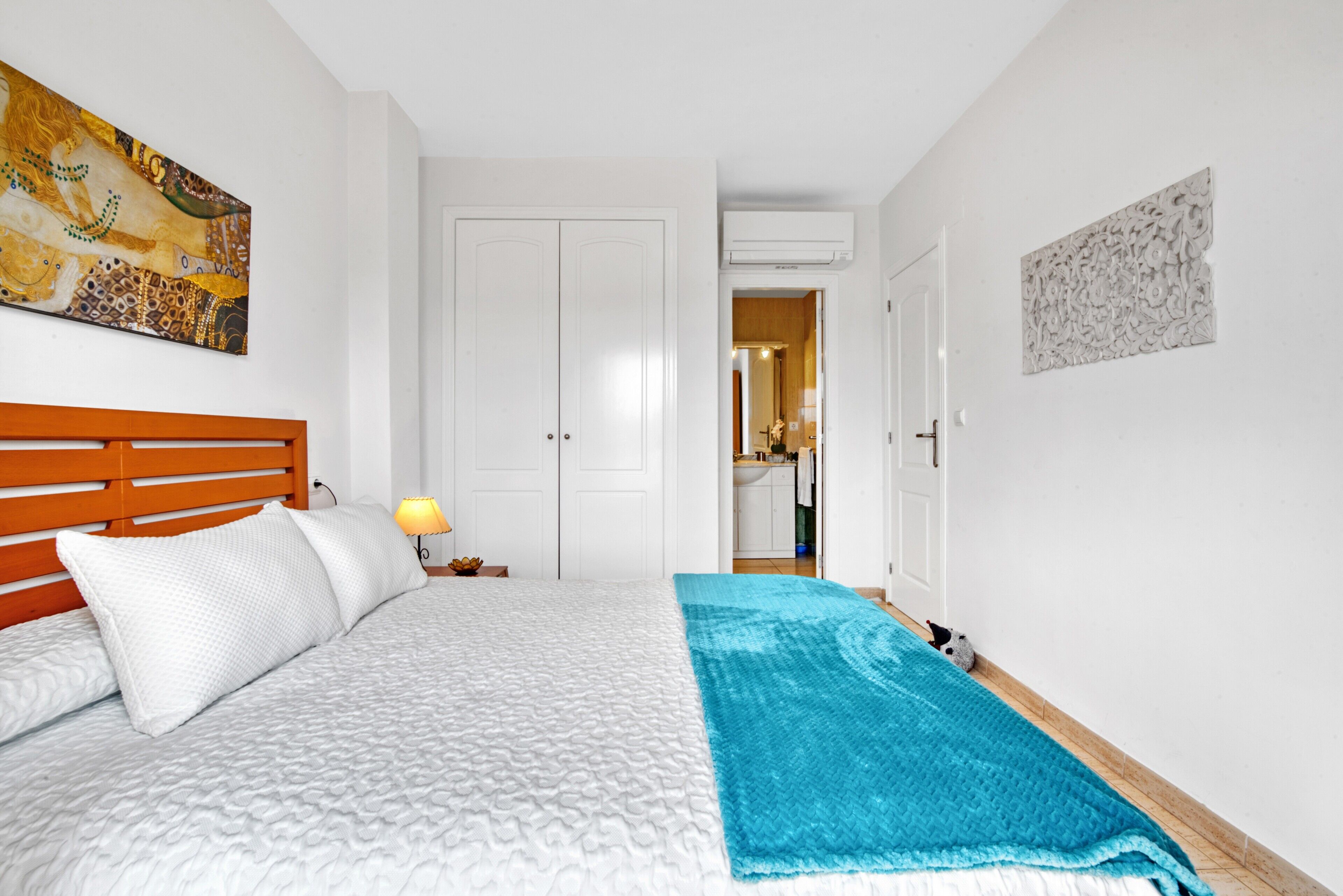 A bedroom with a bed, a wooden headboard, a white wall, a door, and a hallway leading to a kitchen.