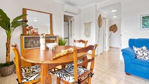 En matplats med ett träbord och stolar, en krukväxt och en blå soffa i ett ljust rum med vita väggar och kakelgolv.