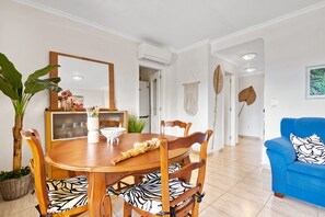 Un comedor con una mesa y sillas de madera, una planta en maceta y un sofá azul en una habitación luminosa con paredes blancas y suelo de baldosas.