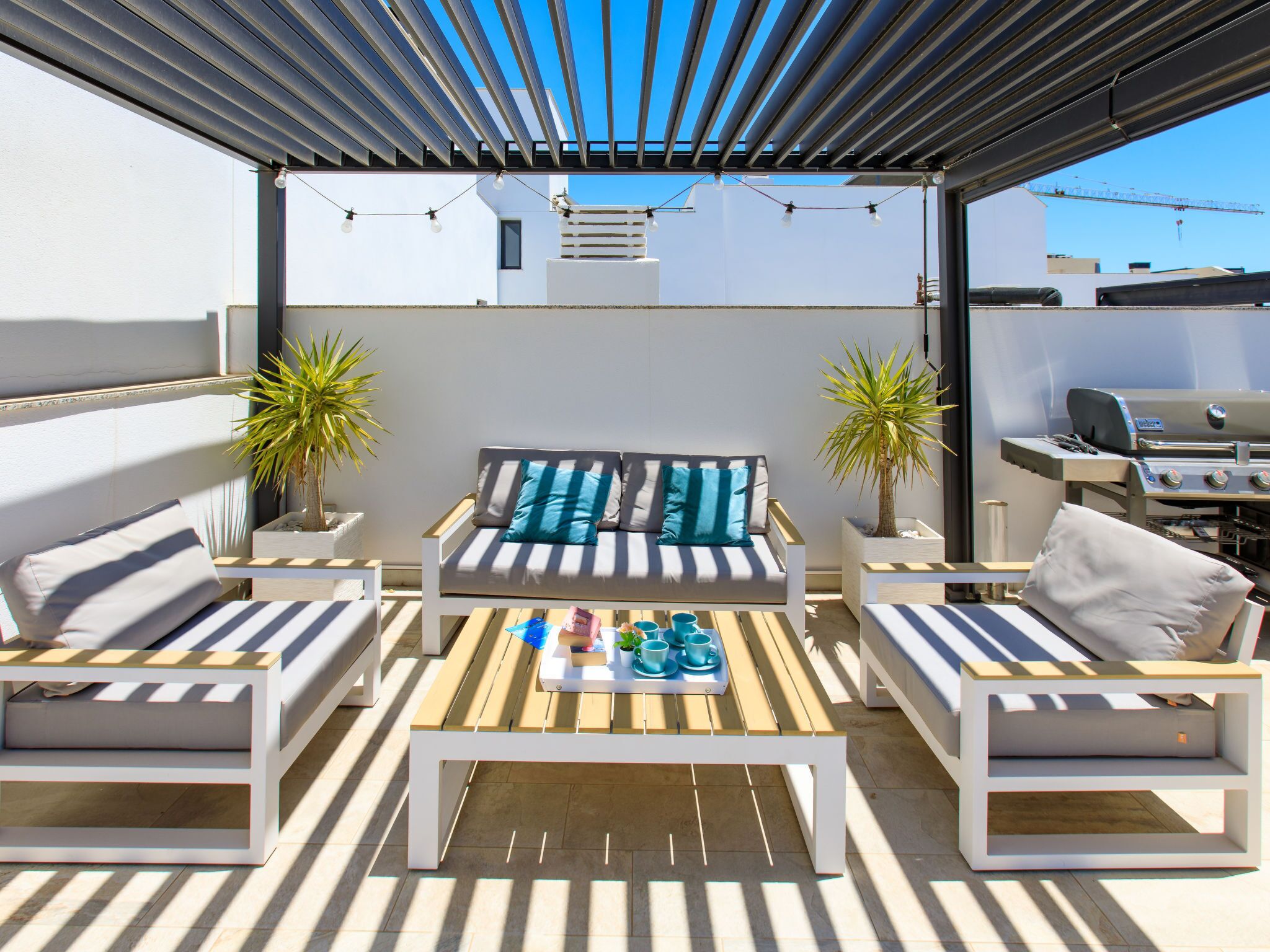 An outdoor patio with a white sofa set, two potted plants, a wooden table with cups and bottles, and a grill.