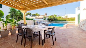 Eine Terrasse im Freien mit einer Holz-Pergola, einem für vier Personen gedeckten Esstisch und einem Swimmingpool im Hintergrund.