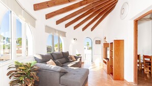 Ein geräumiges Wohnzimmer mit einer grauen Eckcouch, Holzbalken an der Decke und großen Fenstern mit Blick ins Freie.