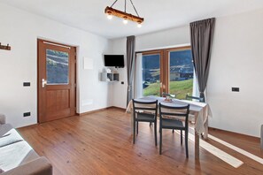 A room with a wooden floor, a dining table with chairs, a TV mounted on the wall, and a window with a view of a landscape. - Apartment 'Val Tort' with Mountain View, Shared Garden and Wi-Fi (Trepalle)