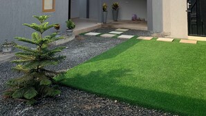 A modern building with a grey facade, large windows, and a black door. There is a small garden with artificial grass and potted plants.