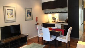 Ein modernes Wohnzimmer mit Flachbildfernseher, Esstisch mit Stühlen und Küchenzeile mit Schränken und Geräten.