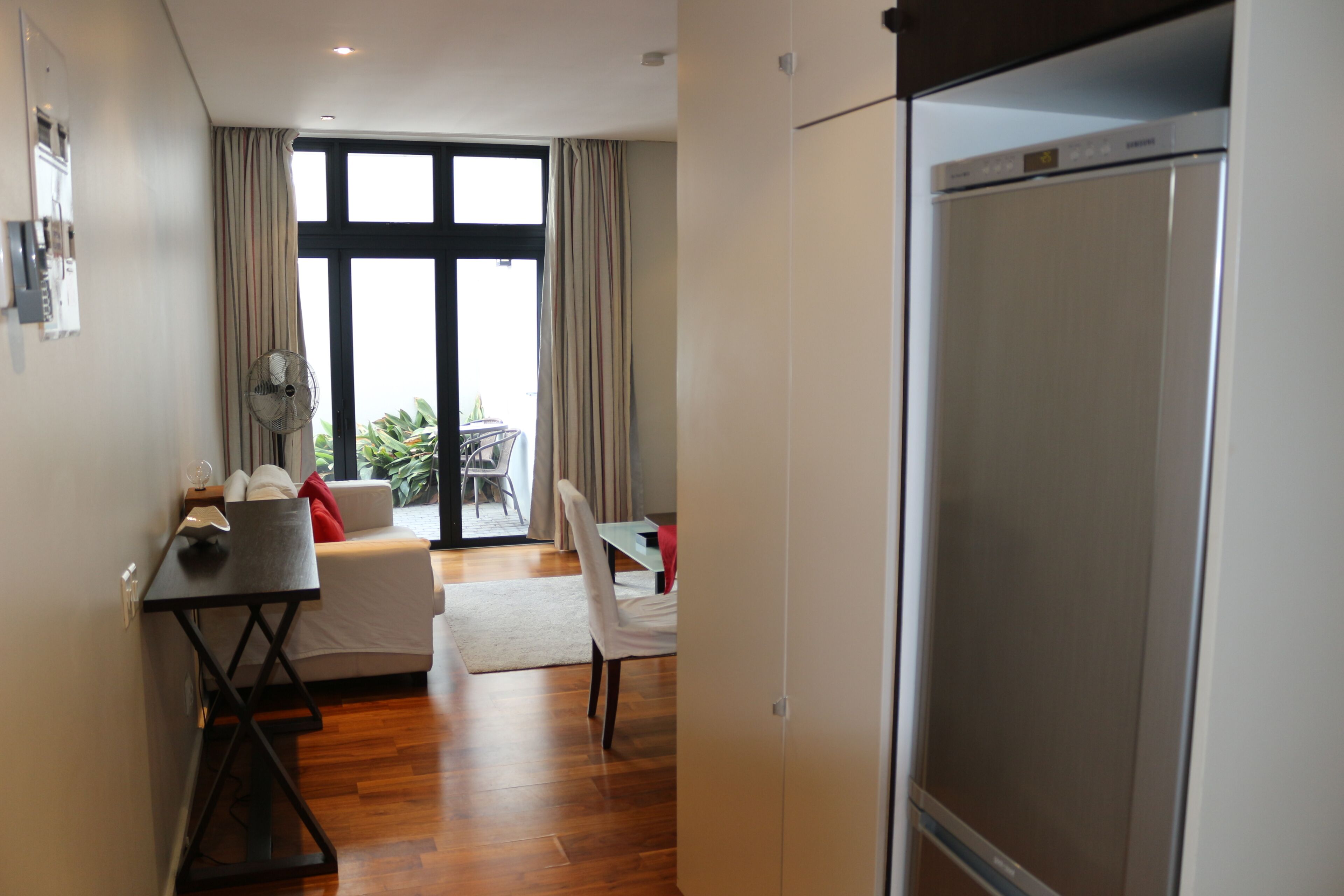 A modern hotel room with a wooden floor, a dining area with a table and chairs, a sofa, and a large window with curtains.