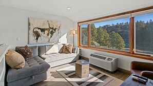 En moderne stue med en grå sofa, et sofabord og et stort vindue med udsigt over et skovområde.