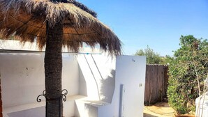 Un patio con suelo de piedra, una estructura de techo de paja, paredes blancas y una cerca de madera.