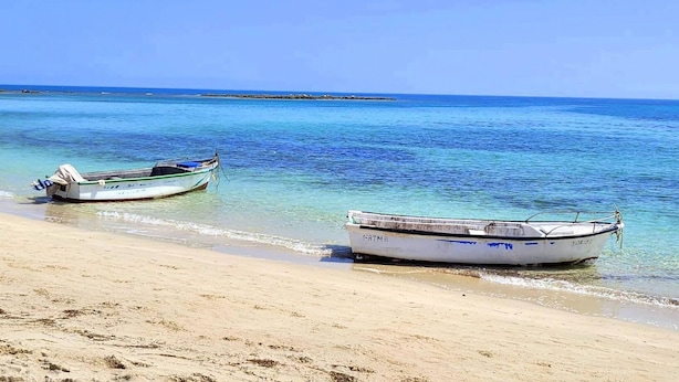 Dos barcos en una playa arenosa con agua azul cristalina y una costa rocosa al fondo.
