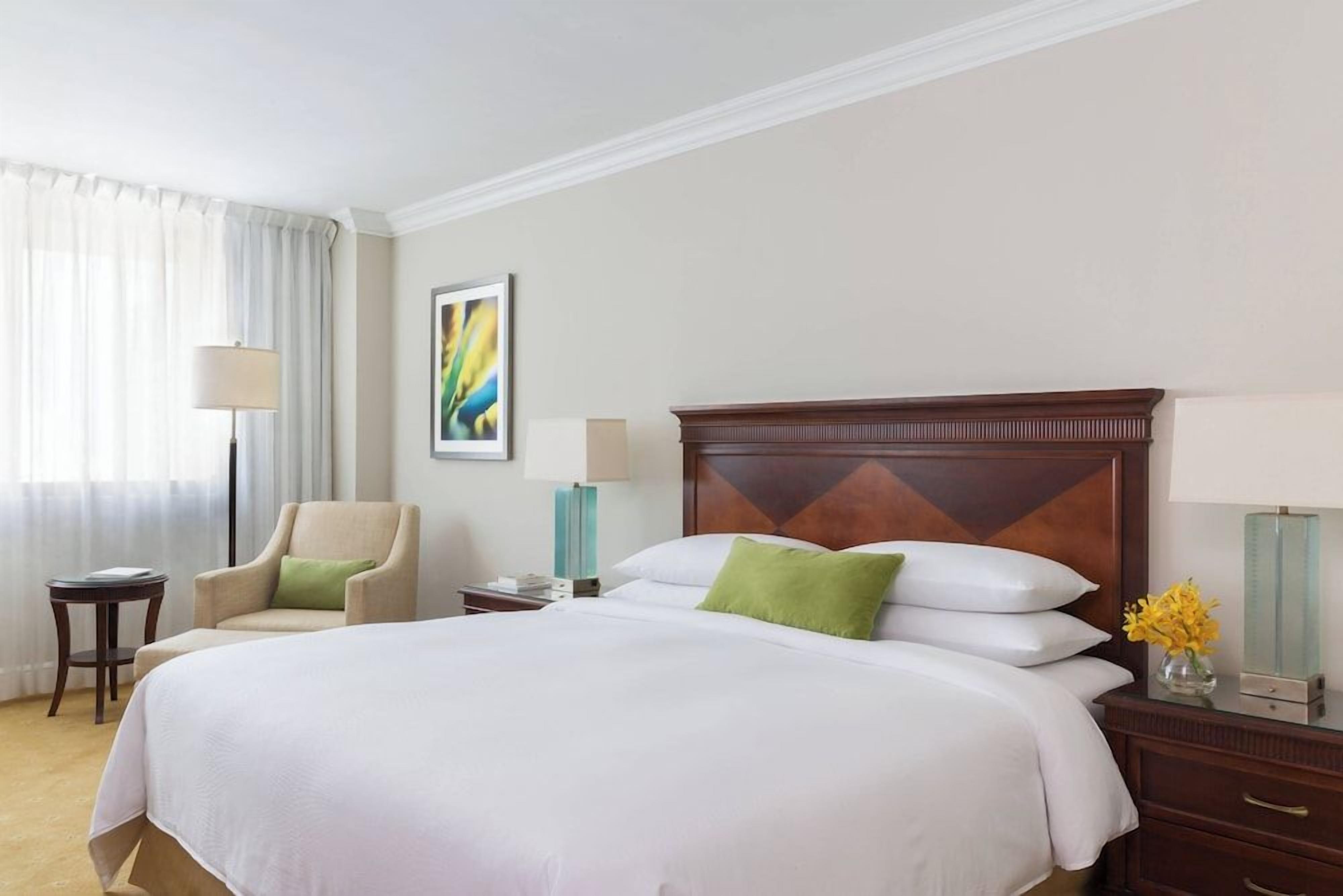 Une chambre d’hôtel avec un grand lit, une chaise, une table de chevet, une lampe et un vase rempli de fleurs.