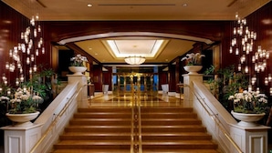A grand staircase with a glass balustrade, leading to a reception area with a chandelier and potted plants.