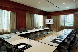 Un salón de conferencias con una larga mesa, sillas, una pantalla de televisión y ventanas con cortinas.