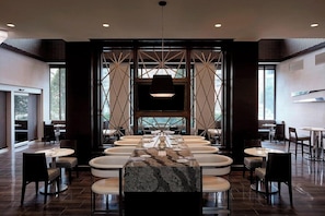 A modern dining area with a long table and multiple chairs, a central decorative light fixture, and a view through a geometric patterned wall. - 2 Connecting Suites at a 4⭐️ Hotel (Arlington)