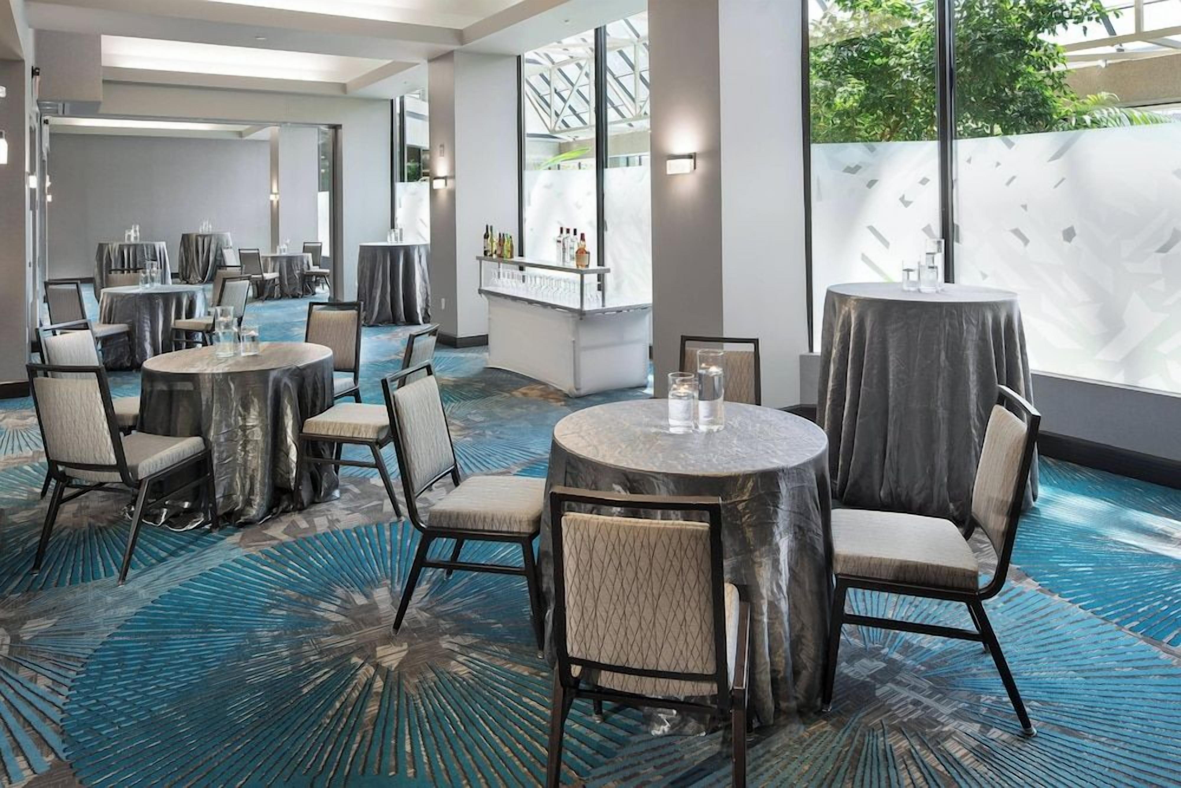 Ein Speisesaal eines Hotels mit runden Tischen, Stühlen, einer Bar und großen Fenstern.