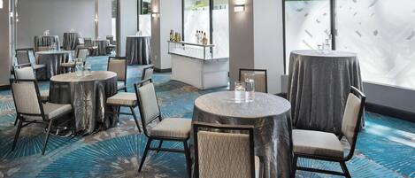 Ein Speisesaal eines Hotels mit runden Tischen, Stühlen, einer Bar und großen Fenstern.