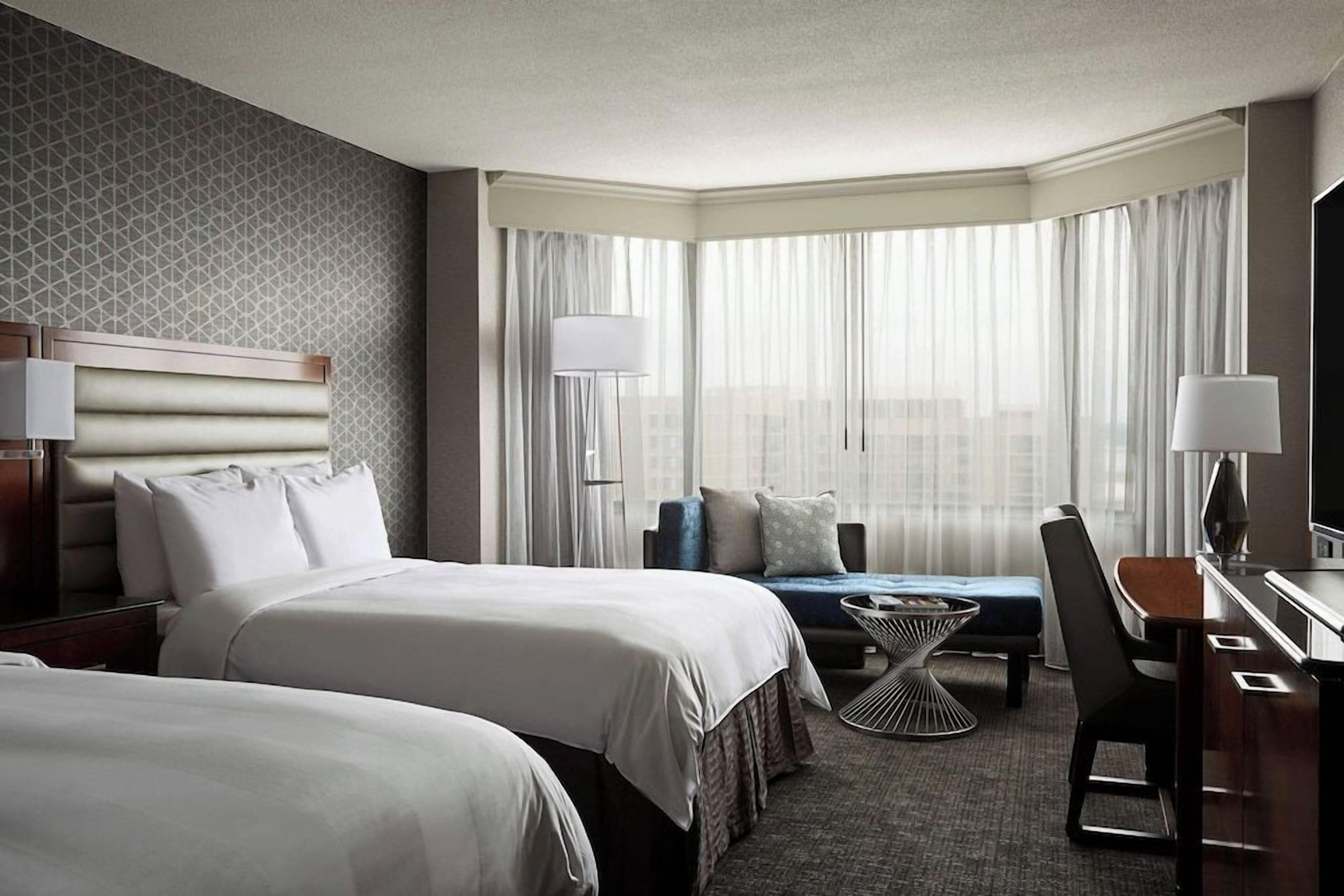 A hotel room with two beds, a desk, a chair, a small table, and a large window with curtains.