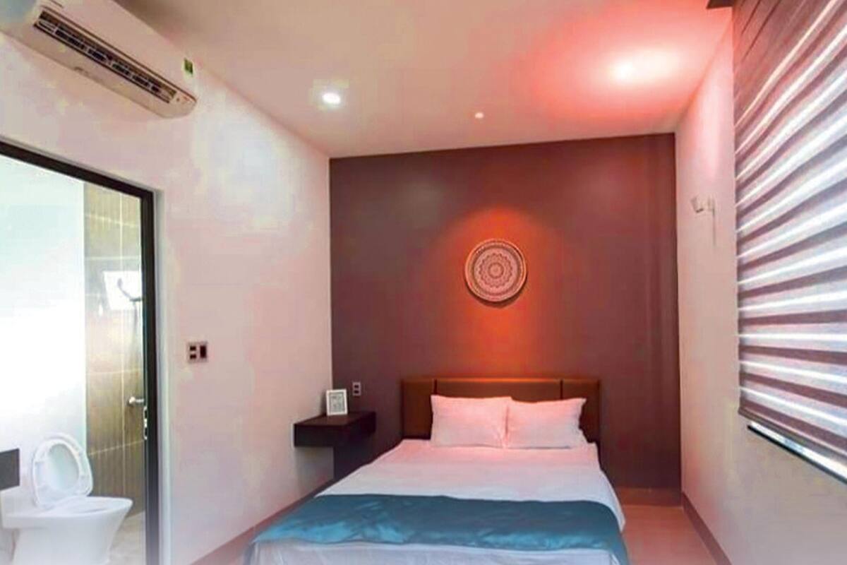 Habitación de hotel con cama, mesita de noche, aire acondicionado de pared y ventana con persianas.