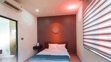 A hotel room with a bed, a bedside table, a wall-mounted air conditioner, and a window with blinds.