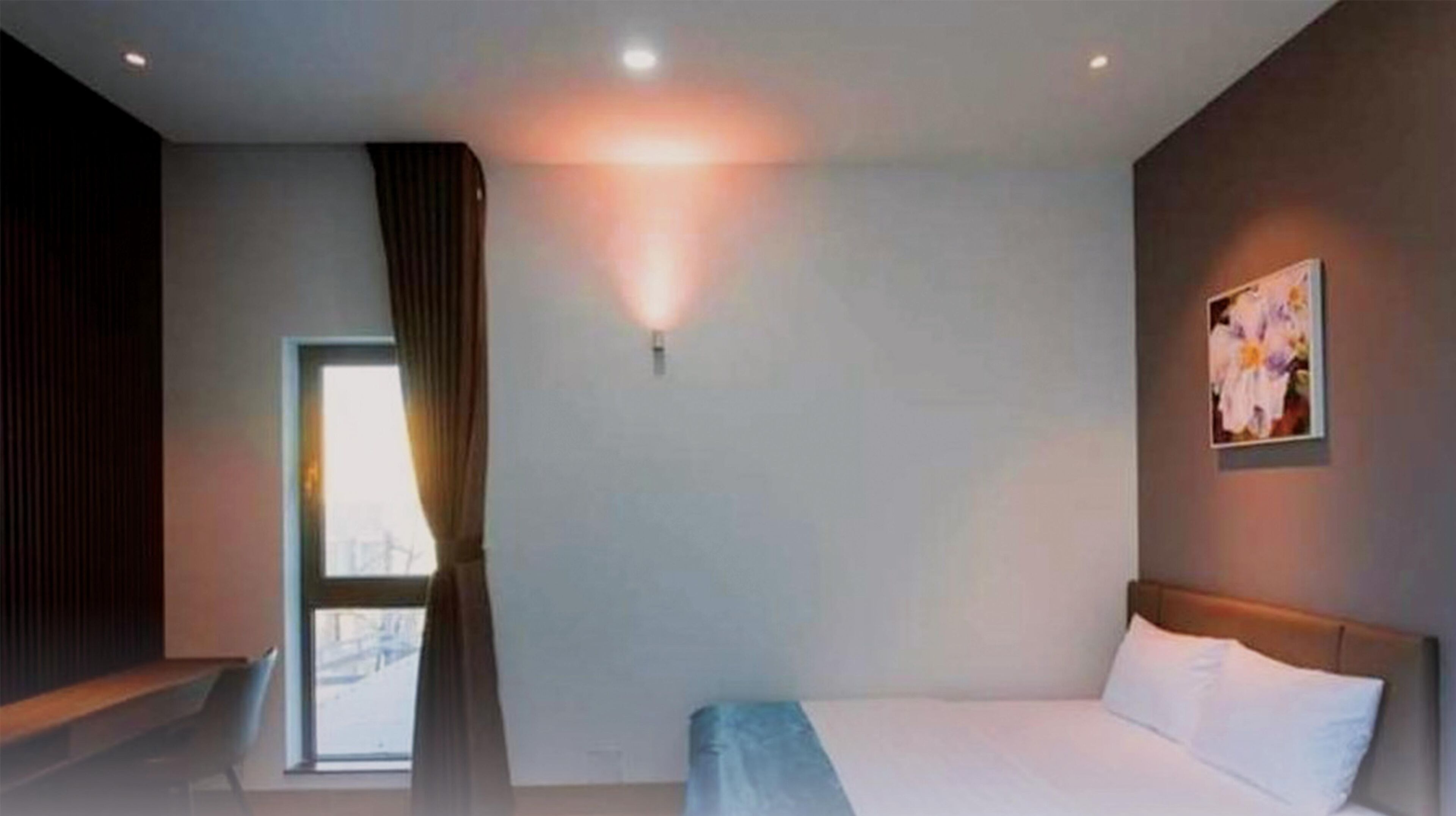 A modern hotel room with a large bed, a window with curtains, a wall-mounted light, and a painting.