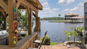 Une terrasse en bois avec un barbecue et des chaises, donnant sur un lac, avec une maison et un quai en arrière-plan.