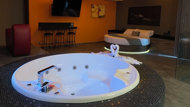 A modern hotel room with a large oval bathtub, a flat-screen TV, a sofa, and a dining area with bar stools.