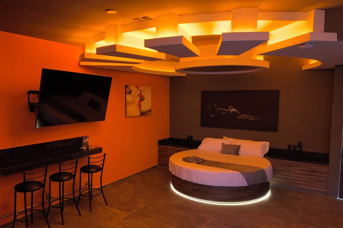 Un dormitorio moderno con una cama curvada, un televisor montado y una zona de bar con taburetes.