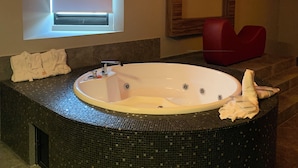 A modern bathroom with a large freestanding bathtub, a window with blinds, a red chair, and a wall-mounted mirror.