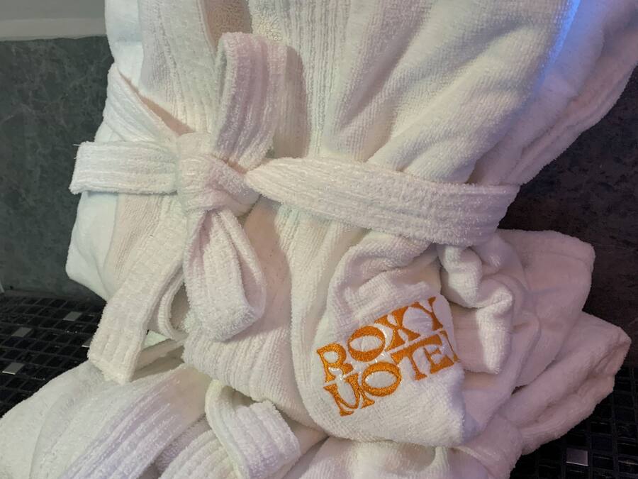 Dos batas blancas con el nombre 'Bobby's' bordado en naranja en las mangas, sujetadas con un lazo blanco, sobre una superficie de azulejos.