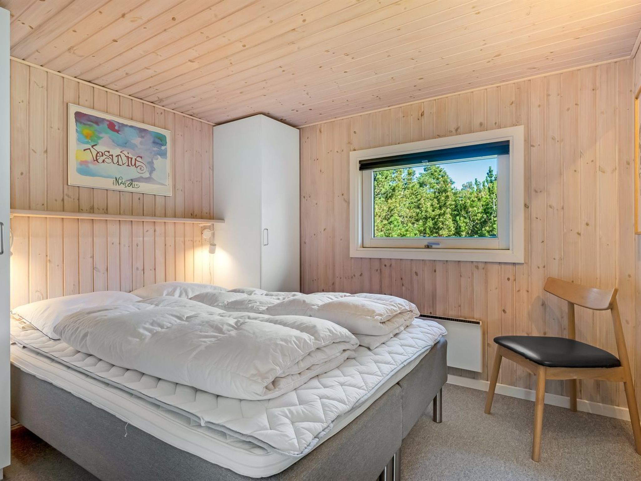Een slaapkamer met een bed, een raam met uitzicht op bomen, een stoel en een kunstwerk aan de muur.