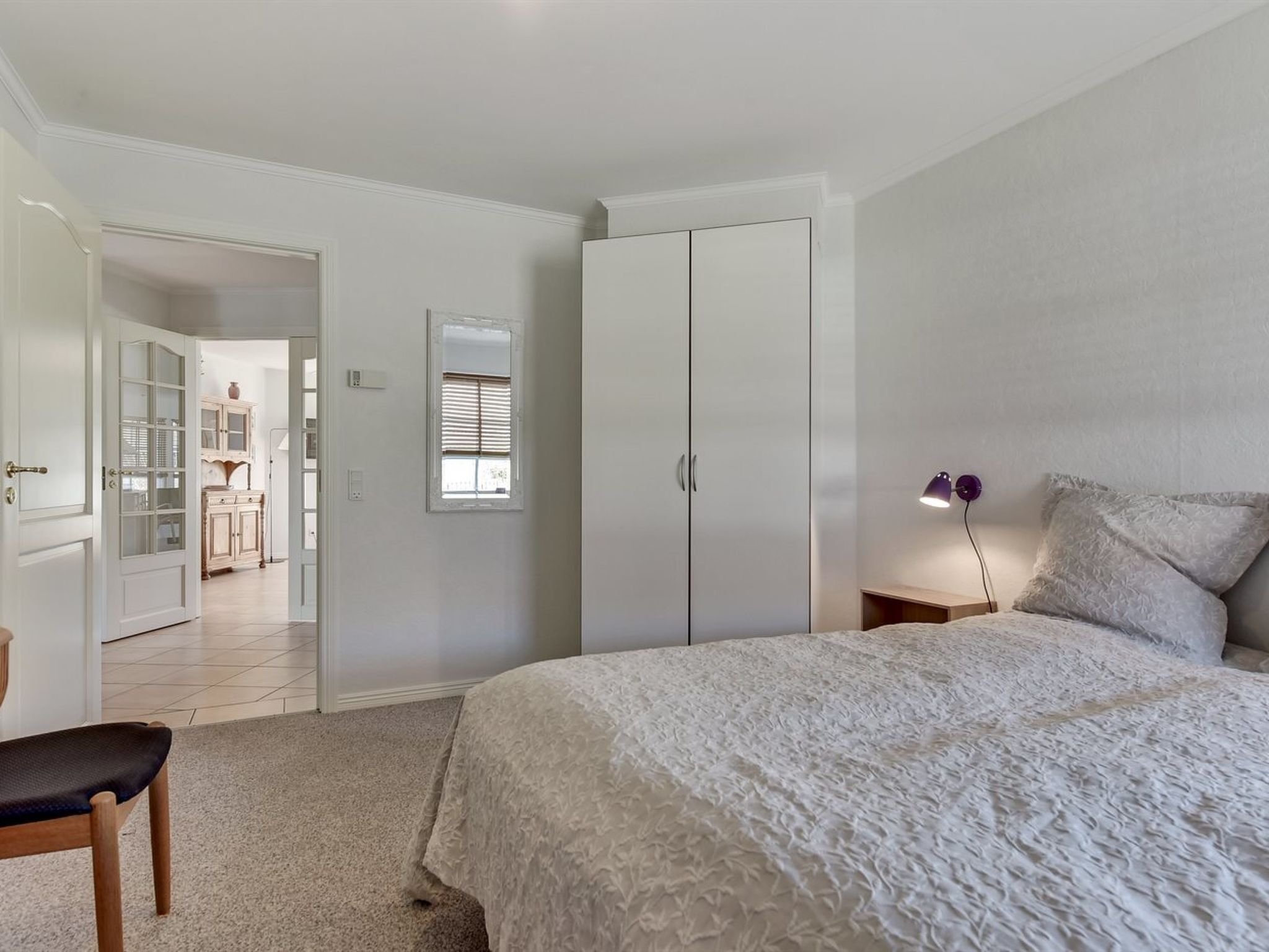 Un dormitorio con una cama grande, una mesita de noche, una lámpara y un armario. Hay una puerta que conduce a otra habitación con un mueble y un espejo.
