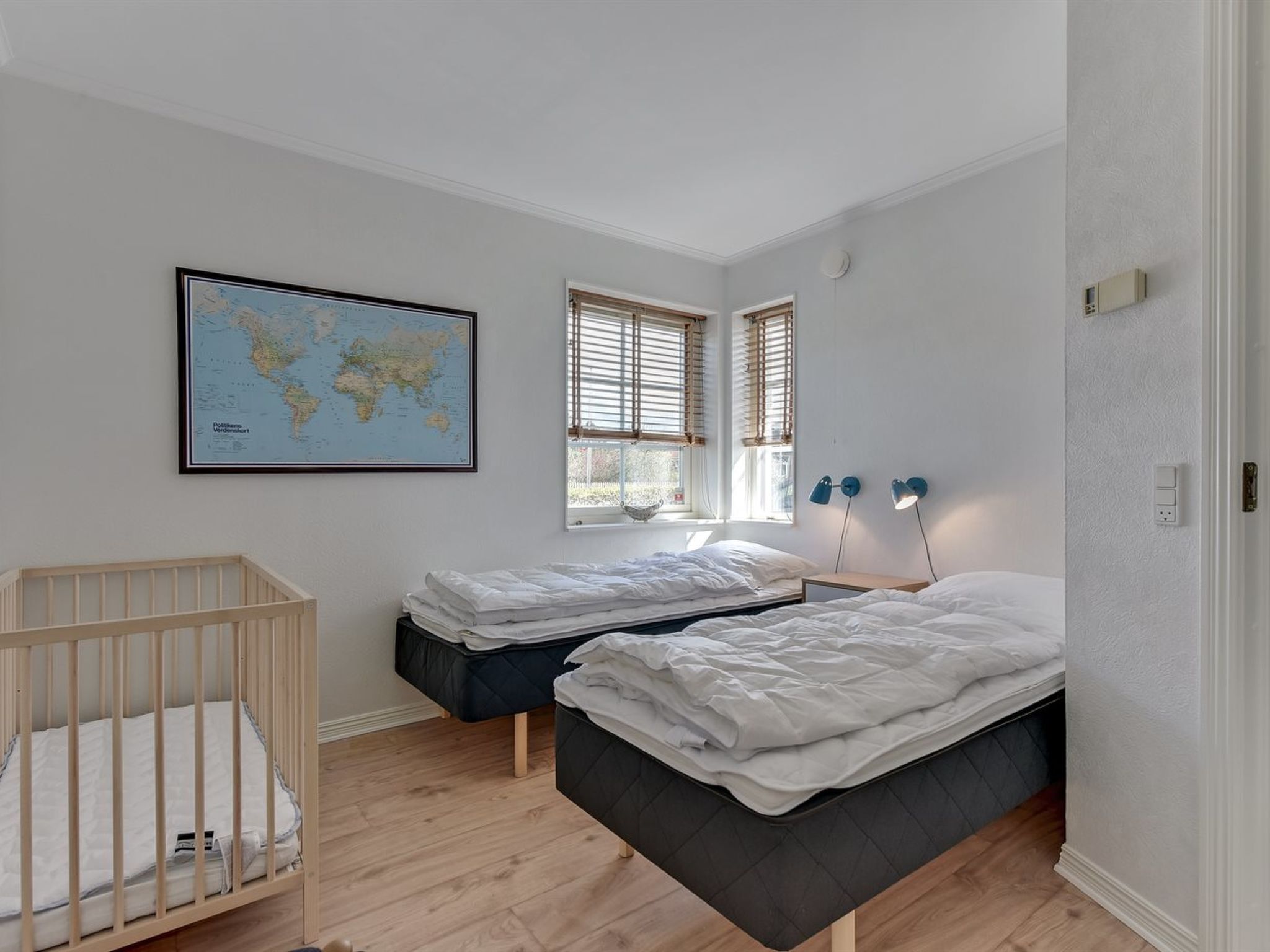 Un dormitorio con dos camas, una cuna, un mapa del mundo en la pared y dos ventanas con persianas.
