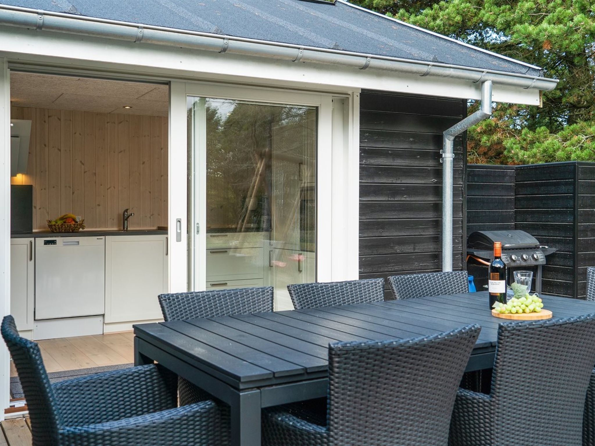 Eine Terrasse mit Tisch, Stühlen, einem Grill und einer Glastür, die zu einer Küche führt.