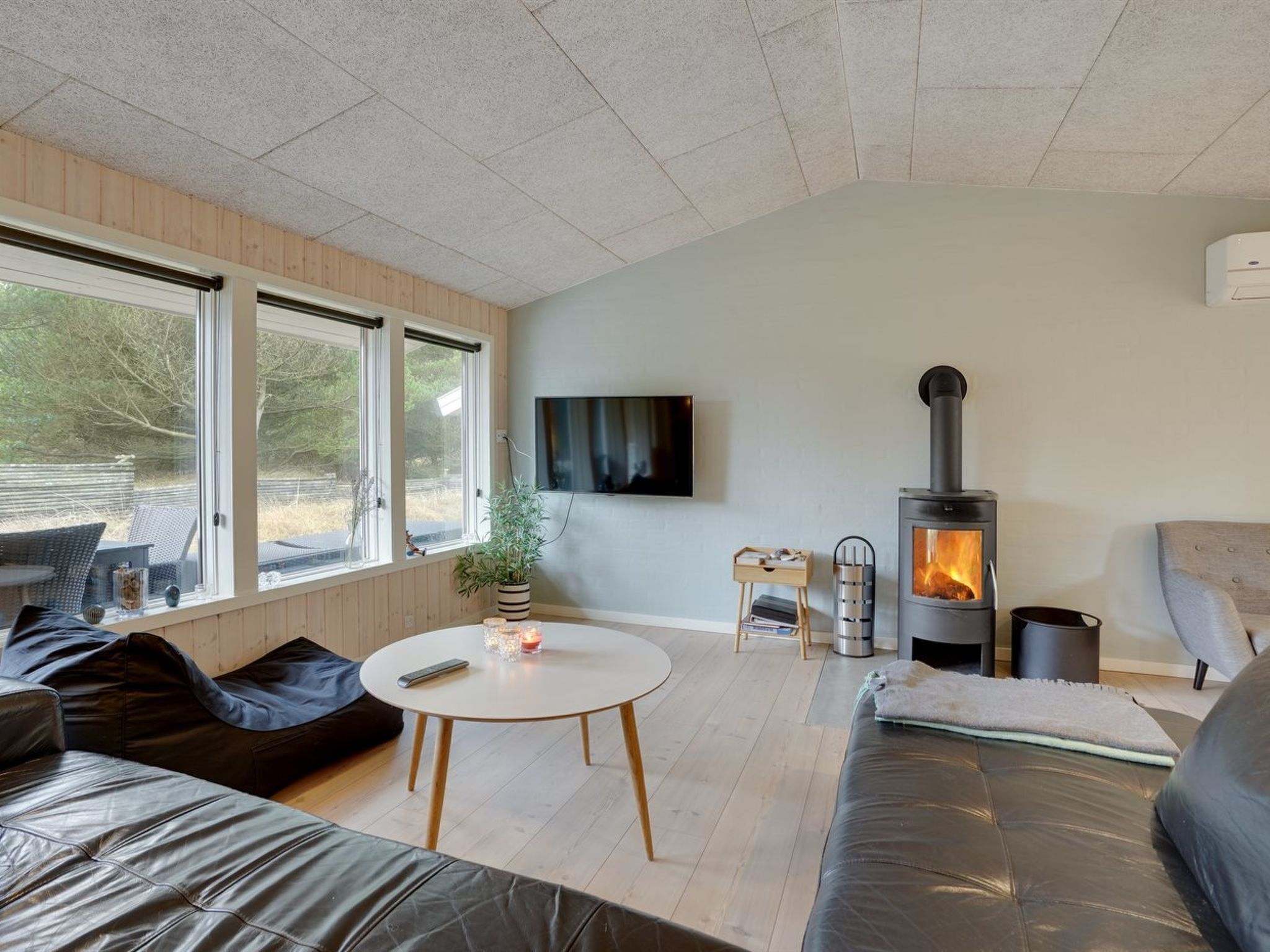 A modern living room with a fireplace, a TV mounted on the wall, and a large window providing a view of the outdoors.