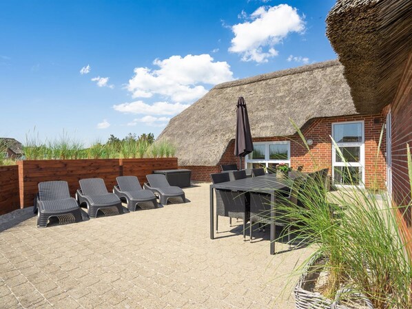 Une terrasse avec des chaises longues et une table, un bâtiment à toit de chaume et un mur en briques.