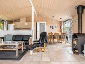 A cozy living room with a sofa, a dining table with chairs, a fireplace, and a kitchen area with wooden walls and pendant lights.