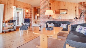 A cozy living room with wooden walls and floors, a brick fireplace, a round wooden table, and comfortable seating. - "Neja" - 400m from the sea by Interhome (Blåvand)