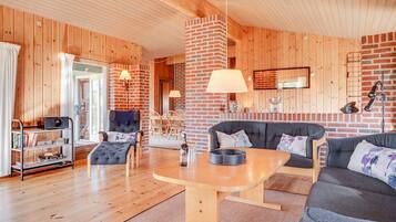 A cozy living room with wooden walls and floors, a brick fireplace, a round wooden table, and comfortable seating.