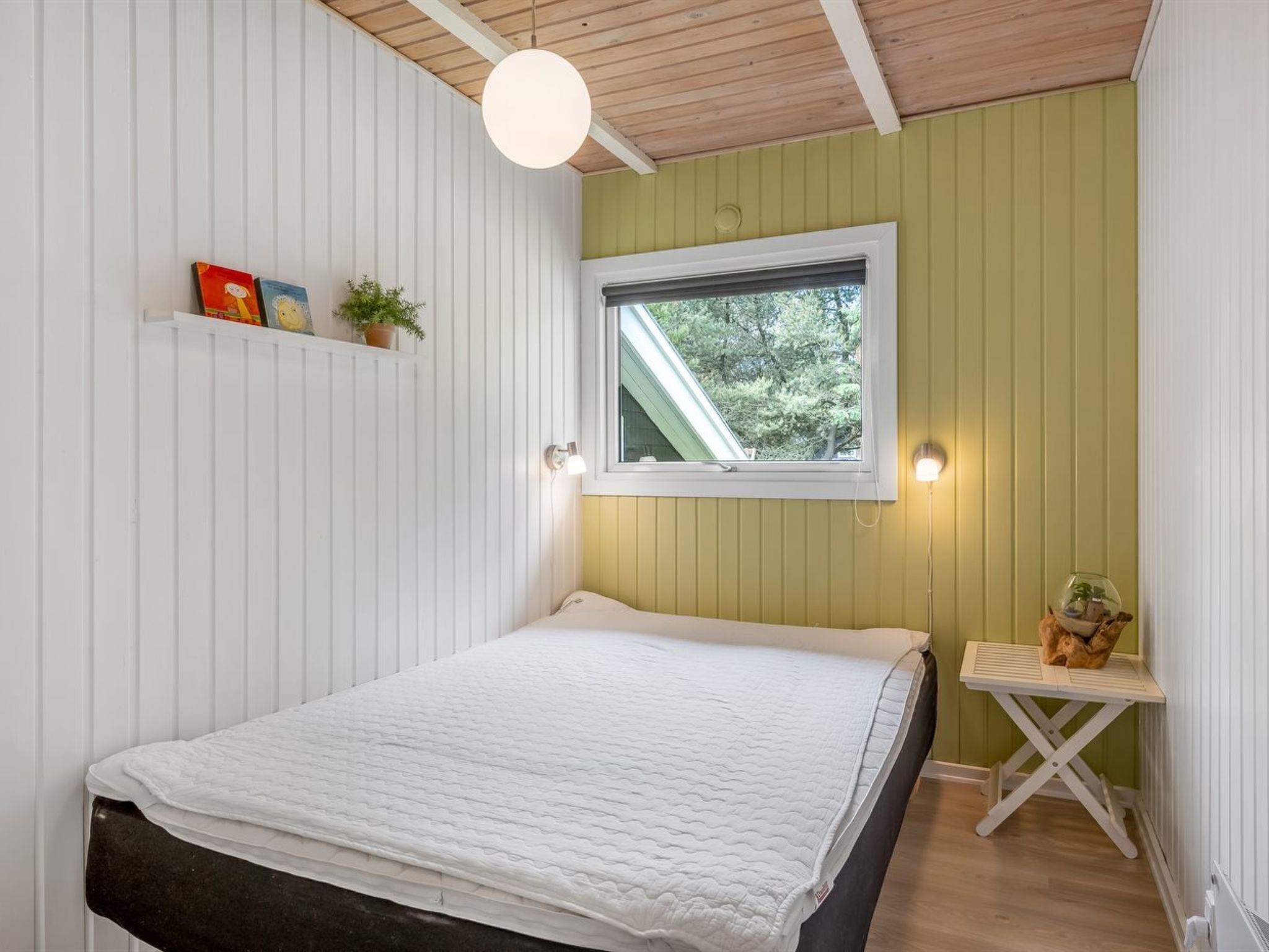 A bedroom with a bed, a window, a shelf with books and a plant, and a small table with a lamp.